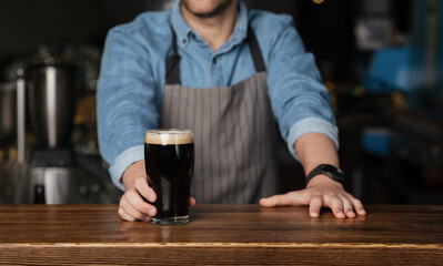 Barman in denim shirt and apron stands behind wooden counter and gives glass of dark beer