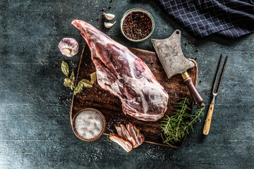 Raw lamb thigh surrounded by salt, pepper, garlic, bacon and rosemary on a vinateg surface