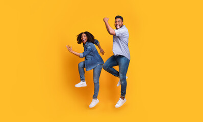 Fototapeta na wymiar Funny African Couple Jumping In The Air And Laughing Over Yellow Background