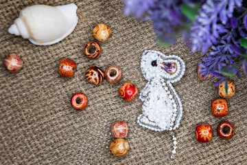 Handmade beaded brooch in the shape of a white mouse. Brooch as a gift and talisman. Handmade jewelry on a fabric background. Selective focus, close-up, composition.