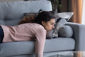 Exhausted young Caucasian woman lying on comfortable sofa in living room sleeping after...