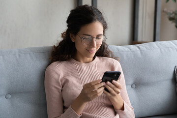 Millennial Caucasian girl in glasses sit relax on couch at home texting or messaging on smartphone, smiling young woman rest on sofa browsing wireless internet, shopping online on web