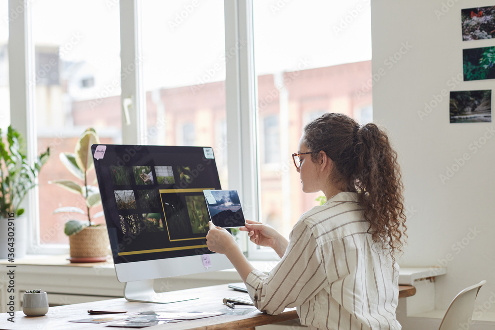 Wall mural back view portrait of modern young woman holding photographs reviewing for publishing while working 