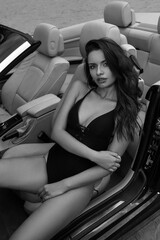 Young sexy female model in black swimsuit. Beaytiful woman sitting in cabriolet at beach. Hot summer day. Fashionable girl with wavy wet hair relaxing in car