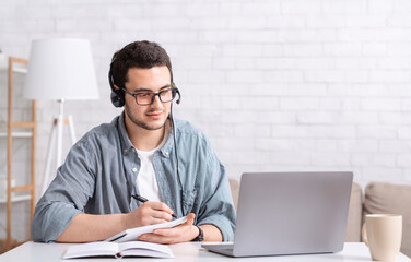Studying and writing in notebook. Busy guy with glasses and headset sitting at table and look at...