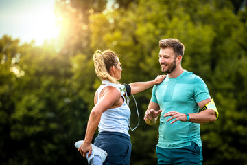 Male personal trainer assisting woman with leg ecercise and talking while smiling before training in park.