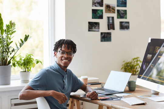Portrait of young African-American photographer smiling at camera while posing at desk with photo editing software on computer screen, copy space
