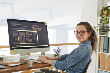 Portrait of young woman looking at camera while writing programming code on computer screen and working at desk in contemporary office interior, copy space - obrazy, fototapety, plakaty