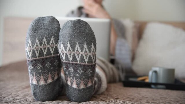 Woman in bed with laptop, close up feet with warm winter socks, drinking hot coffee and eating cookies.