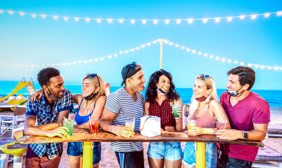 Happy multiracial friends drinking at beach cocktail bar wearing face masks - New normal summer...