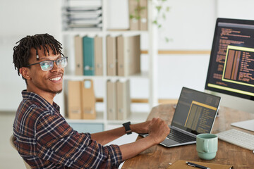 Portrait of smiling African-American IT developer looking at camera while typing on keyboard with...
