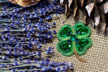 Handmade beaded brooch in the form of a four-leaf clover against a background of dry lavender branches, autumn leaves. A symbol of good luck and wealth in Irish mythology.