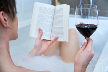 Woman taking bubble bath, reading book and drinking red wine in bathtub. Back view shot.