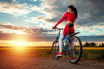 Fototapeta na wymiar Sports training on a bicycle. Beautiful girl in a sports suit on a sunset background. The concept of a healthy lifestyle, cardio training, physical activity. Copy space.