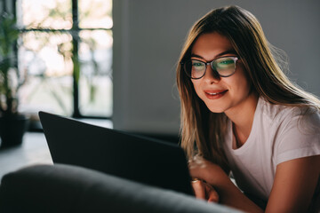 Beautiful young smiling caucasian woman resting on a sofa and using laptop computer. Chatting with friends, browsing information