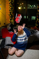 Fototapeta na wymiar A man with a long red beard sits on the floor without pants in a winter sweater. man is holding a wrapped present on the background of New Year decorations and lights. Christmas tree. Parody, humor.
