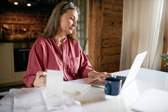 Portrait Of Serious Concentrated Middle Aged Woman Doing Paperwork At Kitchen Table, Having Coffee, Using Online Service To Pay Electricity Bills Or Taxes Over Web, Sitting In Front Of Open Laptop