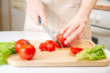 Elegant hands of a young girl cut juicy red tomato into halves on a wooden cutting board. Preparation of ingredients and vegetables. Organic products for diet, nutrition, healthy lifestyle