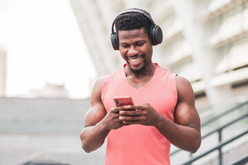 African-American athletic guy listens to music in headphones and smiles in the summer, a man holds a smartphone and looks at the screen