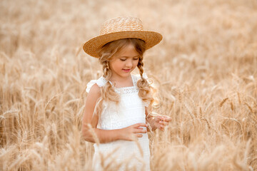 Cute little blonde girl in a straw hat playing in a wheat field on a warm summer day