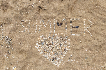 A large, beautiful heart made of sea shells laid out on sea sand with the inscription and letters Summer. Photography, concept, top view.