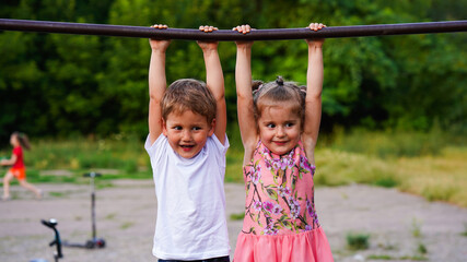 Fototapeta premium Portrait of funny little girl and boy hanging on horizontal bar in summer park