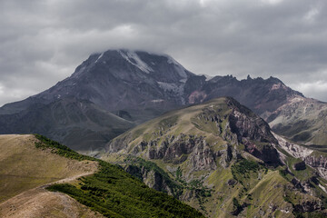 Fototapeta premium Georgia. Caucasus. Green meadows & rock hills on background of Mount Kazbek wrapped by grey clouds