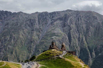 Georgia. Caucasus. Stepantsminda. Trinity Christian Church on background of car parking, group of tourists, green meadows & rock mountain hills with grey cloud sky