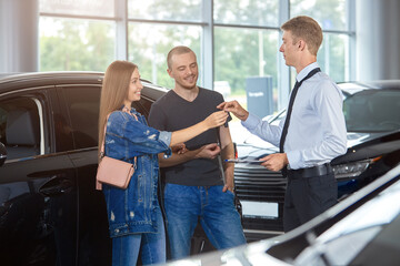 Car dealership Manager congratulates on the purchase of a new car and passes the keys to the couple.