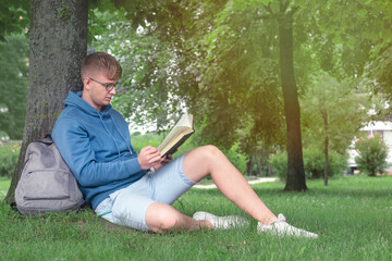 Naklejka na ściany i meble Handsome guy, man, young college or university student or pupil in glasses sitting outdoors in summer park reading book leaning on a tree, learning lessons, preparing for exam in university or college