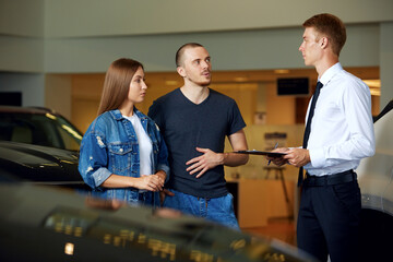 A friendly car dealer shows and explains to a young married couple all the pros and cons of a brand new car at the dealership