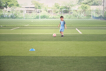 Fototapeta premium Cute little smiling Asian 4 - 5 years old kindergarten boy, football player in soccer uniform is playing football at Training Session, Soccer Drills for Kids concept, Motion blur at football and hand