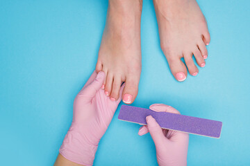 Specialist making french pedicure for client. Closeup young well-groomed female foots. Manicurist doing professional pedicure. Master files nails to client on blue background. Top view.