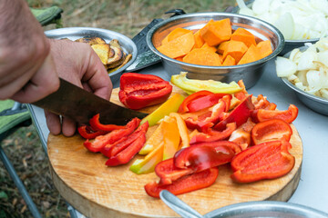 Slicing red and yellow sweet peppers on wooden cutting board. Cooking at the camp.