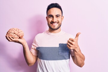 Young handsome man with beard asking for care memory holding brain over pink background smiling...