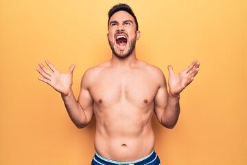 Young handsome man with beard wearing sleeveless t-shirt standing over yellow background crazy and...