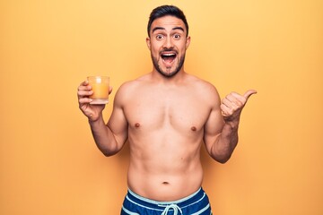 Fototapeta premium Young handsome man with beard on vacation wearing swimwear drinking glass of orange juice pointing thumb up to the side smiling happy with open mouth