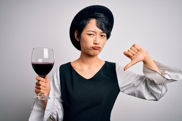 Young beautiful asian sommelier girl drinking glass of red wine over isolated white background with angry face, negative sign showing dislike with thumbs down, rejection concept