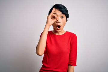 Young beautiful asian girl wearing casual red t-shirt standing over isolated white background doing ok gesture shocked with surprised face, eye looking through fingers. Unbelieving expression.