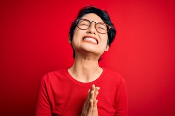 Young beautiful asian girl wearing casual t-shirt and glasses over isolated red background begging and praying with hands together with hope expression on face very emotional and worried. Begging.