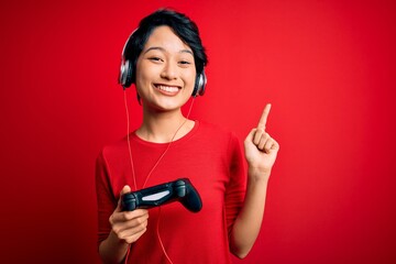 Young beautiful asian gamer girl playing video game using joystick and headphones surprised with an idea or question pointing finger with happy face, number one