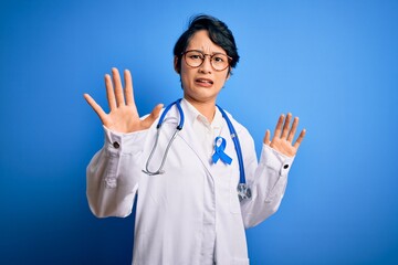 Young beautiful asian doctor girl wearing stethoscope and coat with blue cancer ribbon afraid and terrified with fear expression stop gesture with hands, shouting in shock. Panic concept.