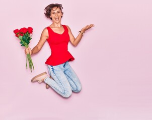Young beautiful woman holding bouquet of roses smiling happy. Jumping with smile on face over isolated pink background.