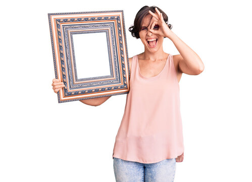Beautiful young woman with short hair holding empty frame smiling happy doing ok sign with hand on eye looking through fingers