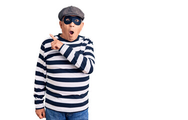 Senior handsome man wearing burglar mask and t-shirt surprised pointing with finger to the side, open mouth amazed expression.