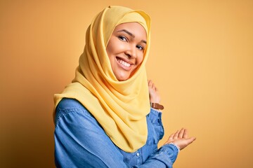 Young beautiful woman with curly hair wearing arab traditional hijab over yellow background Inviting to enter smiling natural with open hand