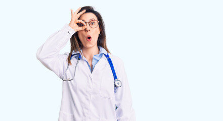 Fototapeta premium Young beautiful woman wearing doctor stethoscope and glasses doing ok gesture shocked with surprised face, eye looking through fingers. unbelieving expression.