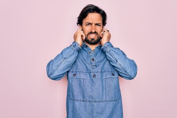 Young handsome hispanic bohemian man wearing hippie style over pink background covering ears with fingers with annoyed expression for the noise of loud music. Deaf concept.