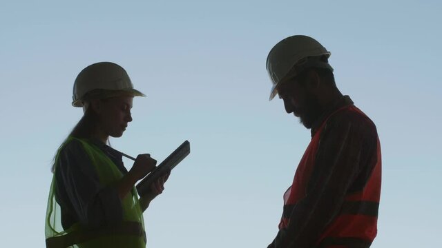 Silhouettes of an engineer with a tablet in his hands and a construction worker in a hard hat against the blue sky stand opposite each other