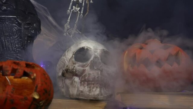 Smoke is blowing at skull, gravestones and pumpkins standing at table. Slow motion of the stream of fume blowing at spooky still life. The conception of halloween and it's scary decorations.